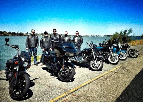 Riders and motorcycles beside the water under a blue sky.