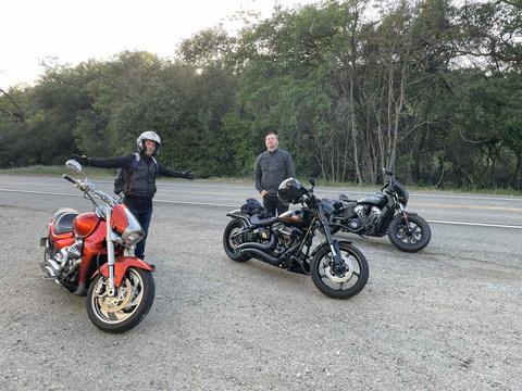 Riders with motorcycles beside a road and green trees.