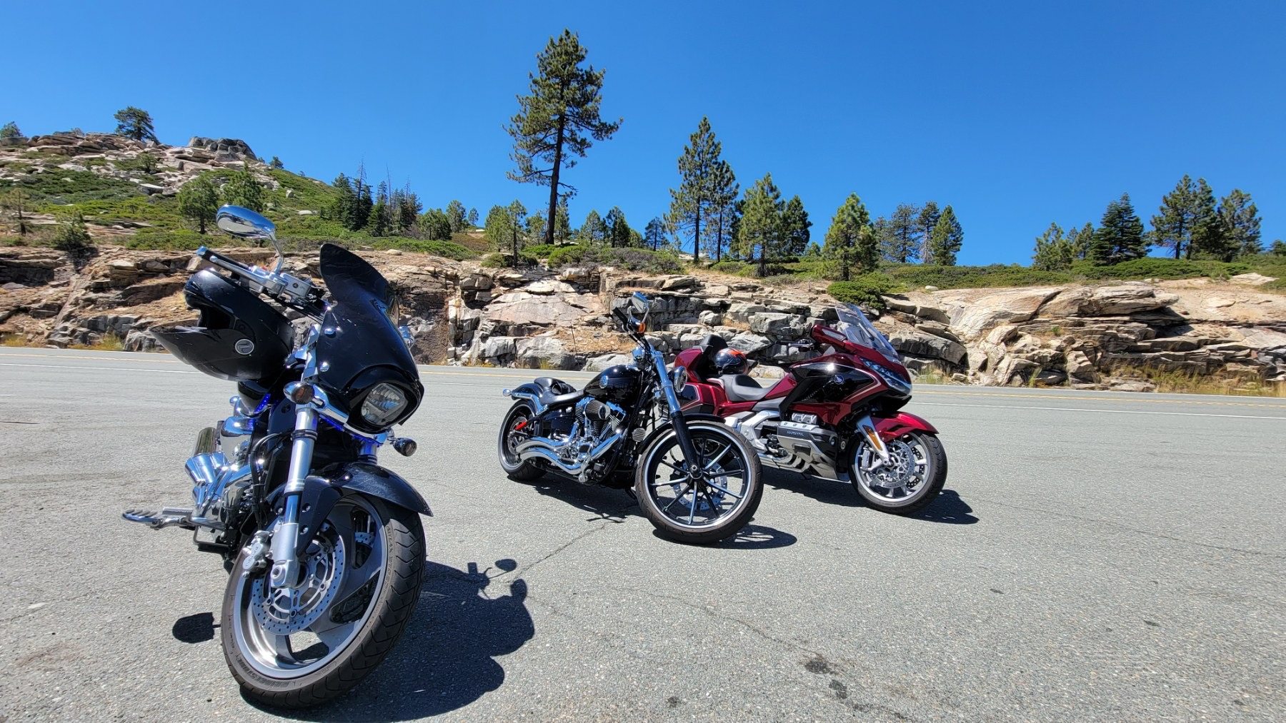 Three motorcycles beside a sunny road with rocky hills behind.