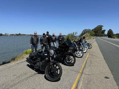 Riders gathered behind several motorcycles beside the water.