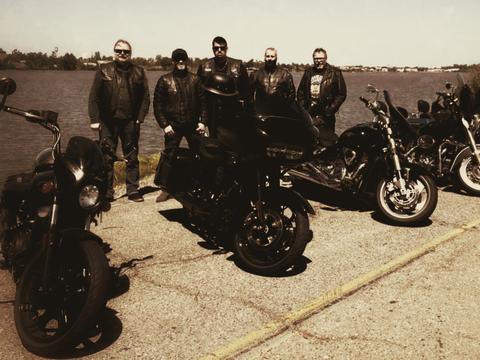Riders standing with motorcycles beside the water in a warm-toned photo.