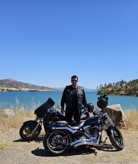 A rider standing beside motorcycles with water and hills behind.