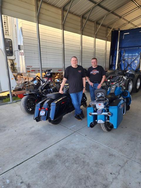 Two people standing beside motorcycles inside a garage.