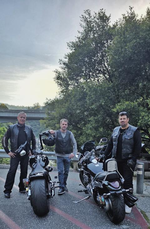 Three riders standing with motorcycles beside a wooded hillside.